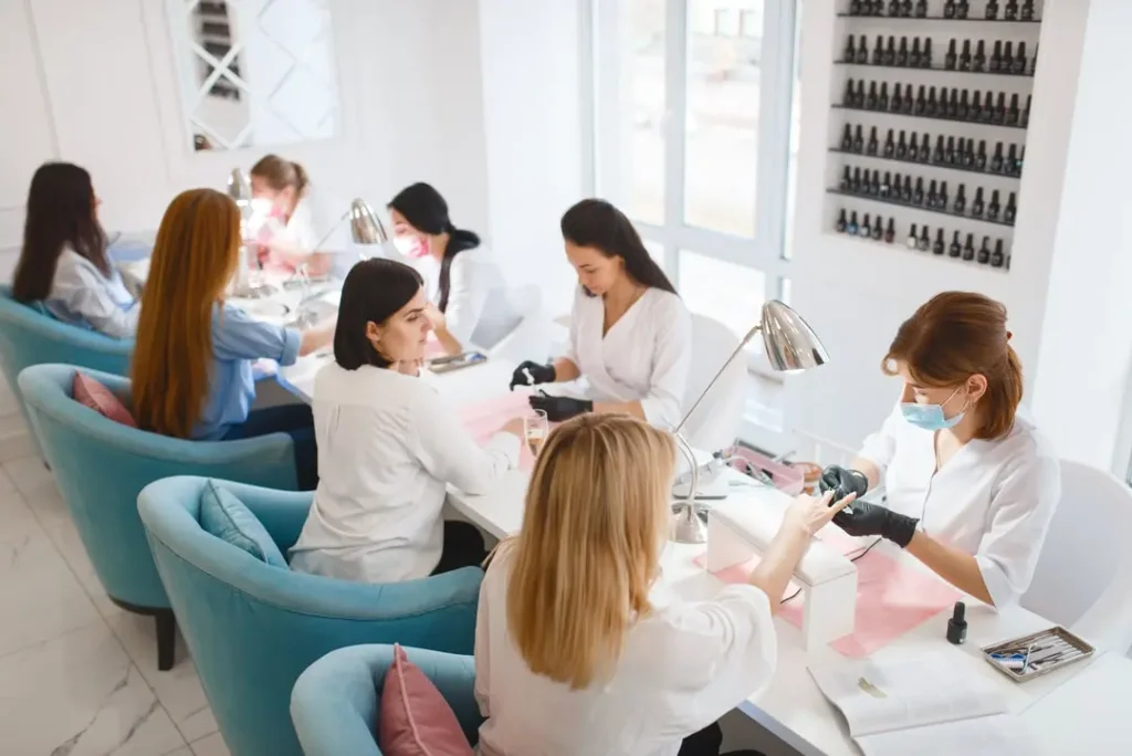 Interior view of a bright nail salon with several technicians providing manicures to clients seated in teal chairs.