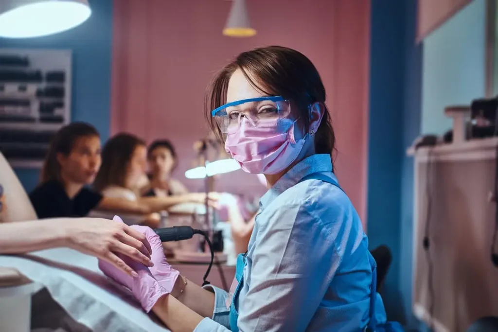 Nail technician wearing mask, safety glasses, and gloves while performing a manicure with an electric file.